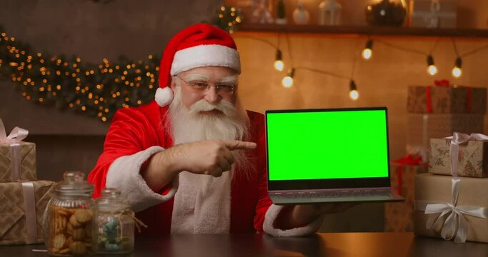 Kind Santa Claus is holding a laptop with a green screen. Indicates the laptop screen. Sits in a beautiful room decorated for a merry christmas.