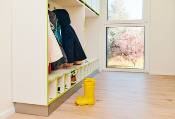 Empty hallway with hangers in day care or kindergarten, yellow rain boots in front of window - obrazy, fototapety, plakaty