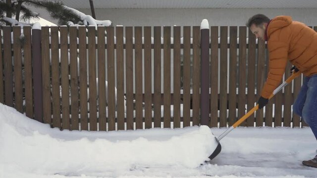 A man shovels snow from the road near the fence with a snow shovel.