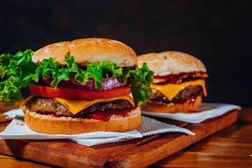 Delicious burgers with Bacon and cheddar cheese and with lettuce, tomato and red onion and bacon and cheddar on homemade bread with seeds and ketchup on a wooden surface and black background.