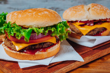 Delicious burgers with Bacon and cheddar cheese and with lettuce, tomato and red onion and bacon and cheddar on homemade bread with seeds and ketchup on a wooden surface and rustic background.