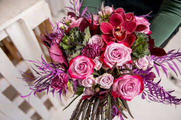 Florist's workplace in a loft space. An experienced florist composes a bouquet of roses, carnations and eucalyptus, adding flowers step by step. She is holding a ready-made bouquet in her hands