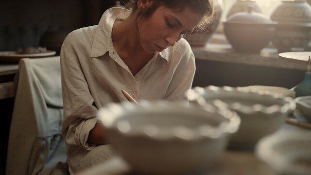 Serious woman scraping clay plate in pottery. Girl carving on product in studio