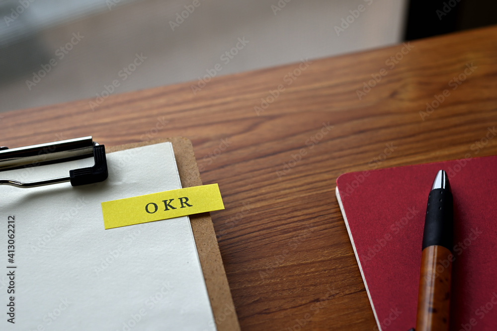Poster there is a notebook, a pen, and a clipboard with a sticky note stuck to it that says okr written on 