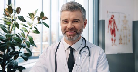 Close up portrait of happy handsome Caucasian middle-aged experienced male doctor in white coat looking at camera and smiling in cabinet in hospital at workplace, man infectionist, healthcare concept