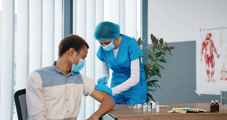 Female young professional healthcare assistant nurse in protective uniform preparing injection and injecting covid-19 vaccine to handsome male patient in medical mask sitting in hospital lab room