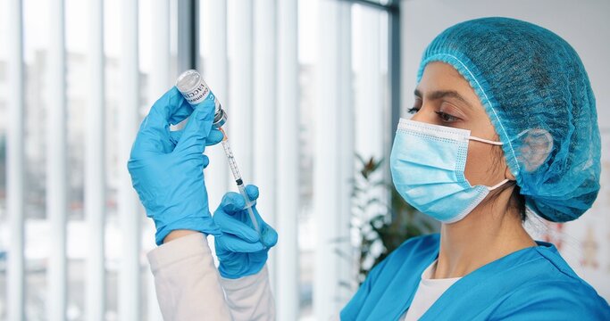 Close up of young busy Hindu female healthcare specialist nurse in protective uniform and medical mask holding in hands ampoule with coronavirus vaccine preparing injection for covid vaccination,