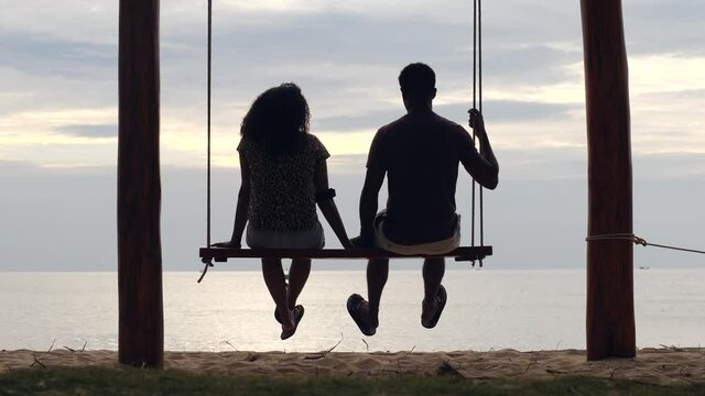 Young couple sitting on a swing at the beach in front of the calm sea and bright sky.
