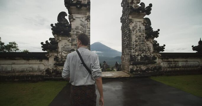 Dolly medium shot of a man walking towards Gates of Heaven temple gates in Pura Penataran Agung Lempuyang temple in Bali with reveal of the Mount Agung volcano and temple grounds below