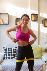 Fototapeta na wymiar Healthy and attractive young woman is exercising in her living room. She wears black leggins, a pink top, and a green league