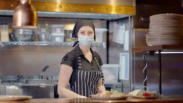 Confident young woman in coronavirus face mask putting plate with salad on counter in slow motion and looking at camera. Middle shot portrait of professional cook working in restaurant on pandemic.