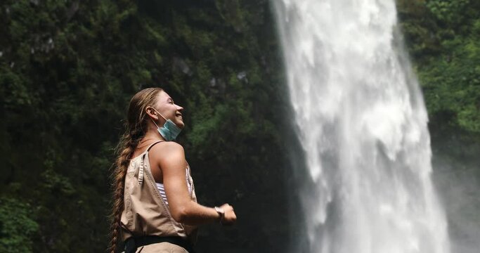 A woman smiling removes a medical mask against the background of a waterfall