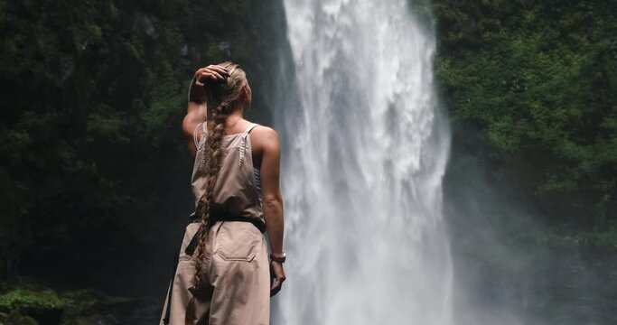 A girl in a jumpsuit enjoys the view of the waterfall adjusting her long hair