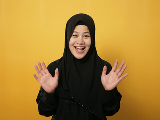 Muslim woman wearing hijab looking at camera smiling and waving her hands saying hi or goodbye farewell gesture against yellow background