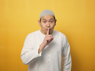 Asian muslim man shushing gesture, put his finger on lips telling secretly gossip concept, against yellow background