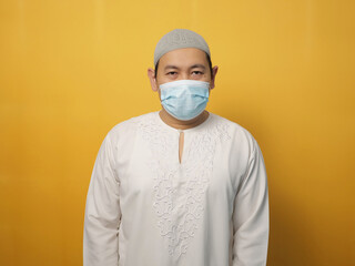 Portrait of Asian muslim man wearing facial mask, against yellow background, new normal due to covid coronavirus pandemic