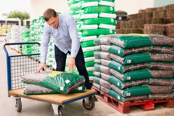 Glad cheerful positive male buyer storing fertilizer bags on trolley in hardware store