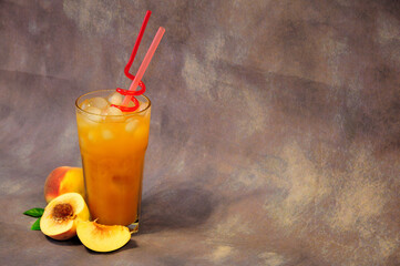 Tall glass of fruit nectar with ice and slices of ripe peach next to it on a gray background.