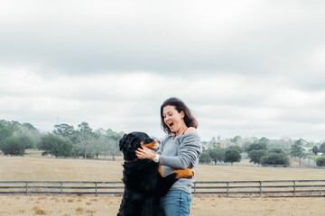 Happy woman with active dog playing outdoor. Cheerful owner and big bernese mountain dog have fun on field against village landscape. Walking with pet.