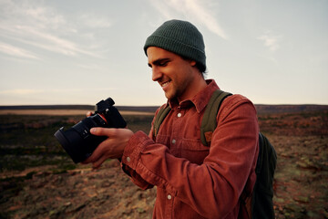 Male travel photographer checking photos on camera after shot standing on mountain wearing cap and...