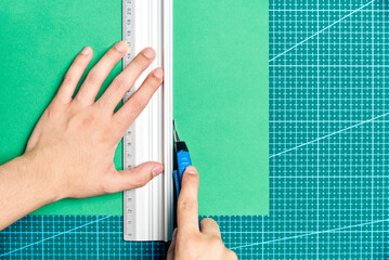Hands cutting a green sheet of paper with a cutter and a metal ruler on a cutting board