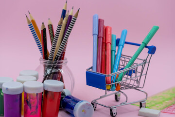 Bright stationery objects in mini supermarket cart on pink background. Back to school concept.