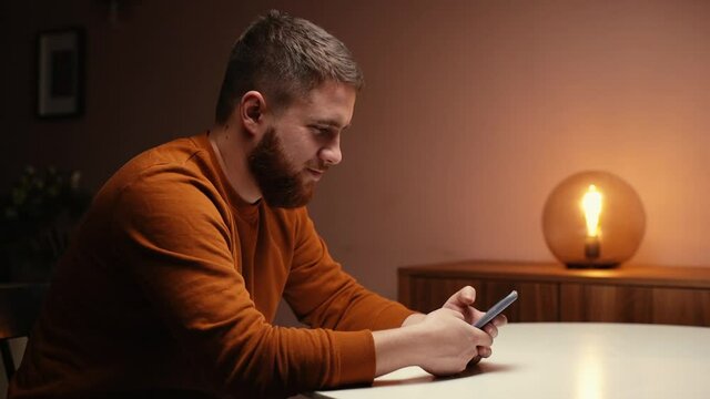 Side view of bearded young man using mobile phone sitting at table in dark living room. Cheerful male typing message on smartphone at home. Smiling business man having break with cell phone.