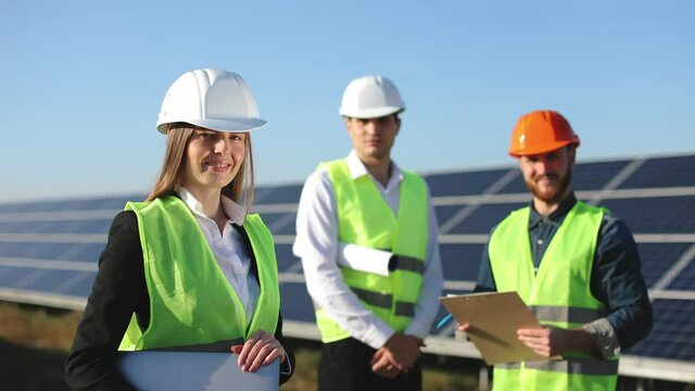 The female engineer is standing and looking at the camera. She is smiling. Two engineers are also looking at the camera. They are standing next to solar panels. 4K.