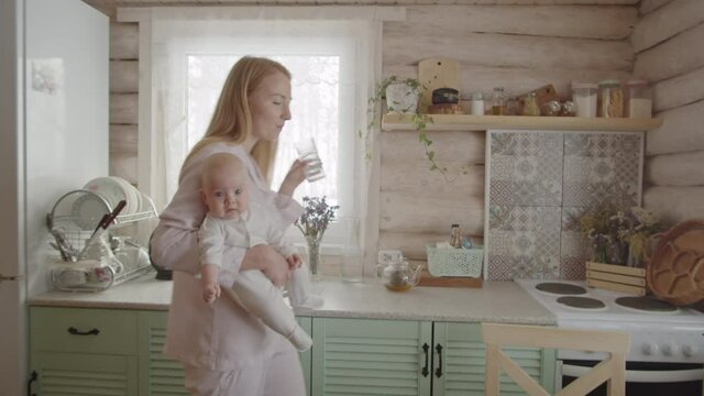 Panning shot of young mother in pajamas holding baby daughter, drinking water and then sitting at kitchen table and using laptop