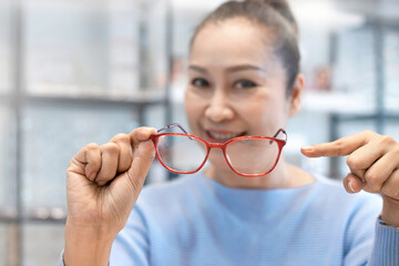 Cheerful middle age Asian woman is testing a new spectacles that she just received from her Optimist  in optical store, choosing and picking frame, eyesight and vision concept, selective focus