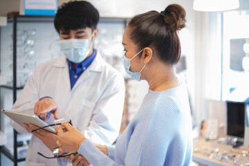 Fototapeta premium Asian optometrist in white coat and medical mask gives senior woman advice on new model of glasses