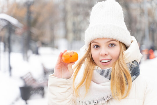 cute caucasian teenage girl in white warm clothes shows the emotion of surprise and joy in winter holding a tangerine with a painted face