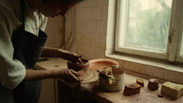 Focused master making new clay product in pottery. Woman scraping line on pot 