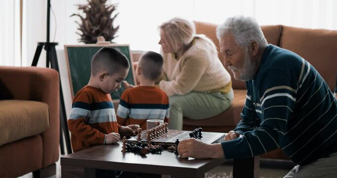 Grandfather shows the grandson how to play chess, grandmother and grandson draw in the background on the board at home. Twin boys with grandparents, free days for family fun
