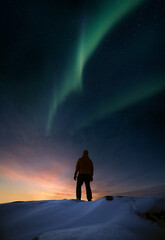 A person standing on snowy rock in winter and looking at sunset and aurora borealis - 413147023