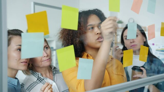 Multi-ethnic creative team of women discussing business writing on colorful sticky notes on glass board working indoors in modern office. People and creativity concept.
