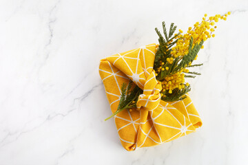 Top view of wrapped gift by linen fabric in furoshiki eco style with yellow mimosa flower. Woman or mother day celebration concept.