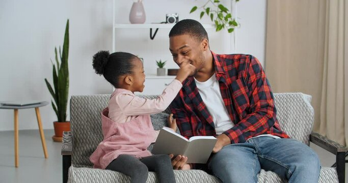 Young dad reads book to his beloved daughter shows pictures explains talks with child sitting on sofa in living room, African American man parent teaches little girl to read, family laughs at home