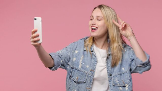 Funny blonde young woman 20s in white t-shirt denim jacket isolated on pink background studio. People lifestyle concept. Doing selfie shot on mobile phone blowing sending air kiss showing victory sign
