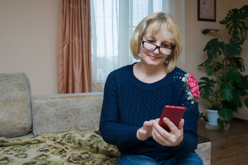 A beautiful blonde woman 50-60 years old with a phone in her hands sits on the couch at home. Grandma is making a video call. Affordable modern technologies for retirees. Place for text