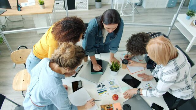 Cheerful and hard-working employees discussing business ideas working in modern office standing around tables with graphs and charts. Teamwork and job concept.