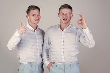 Happy men together. Two brothers showing thumbs up and ok gestures. Models winking and smiling on grey background. Twins wearing blue shirts. Brotherhood and friendship concept.