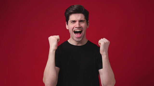 Surprised shocked young man 20s years old in casual black t-shirt posing isolated on red color background in studio. People lifestyle concept. Say wow spreading hands screaming doing winner gesture