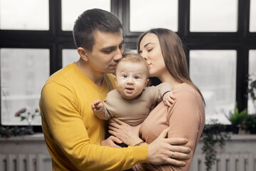 Happy family parents kiss small baby son on background of window, sunlight