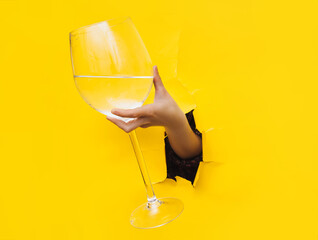 A left woman's hand emerges through a torn hole in light yellow paper with a large glass with vermouth, water or vodka. The concept of alcoholism, drunkenness and hangover. Copy space.
