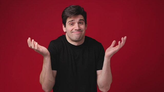 Confused puzzled young man 20s years old wearing casual basic black t-shirt posing isolated on bright red color background in studio. People lifestyle concept. Spreading hands say oops looking camera