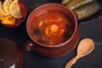Homemade Traditional Russian soup Solyanka with meat, sausages, vegetables, pickles and olives with lemon in ceramic pot with wooden spoon on dark wooden background