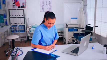 Healthcare assistant taking notes on clipboard and typing on laptop working in hospital clinic. Medical physician in medicine uniform writing list of consulted, diagnosed patients, making research.