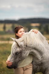 Girl horse rider stands near the white horse and hugs the horse. Horse theme outdoors scene in countryside. Young woman rider and her beautiful white horse