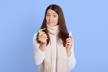 Pretty female wearing casual attire looking at camera with astonished facial expression, holding remedy in hands, using sprays for throat and nose, stands against blue wall, ill lady treating.